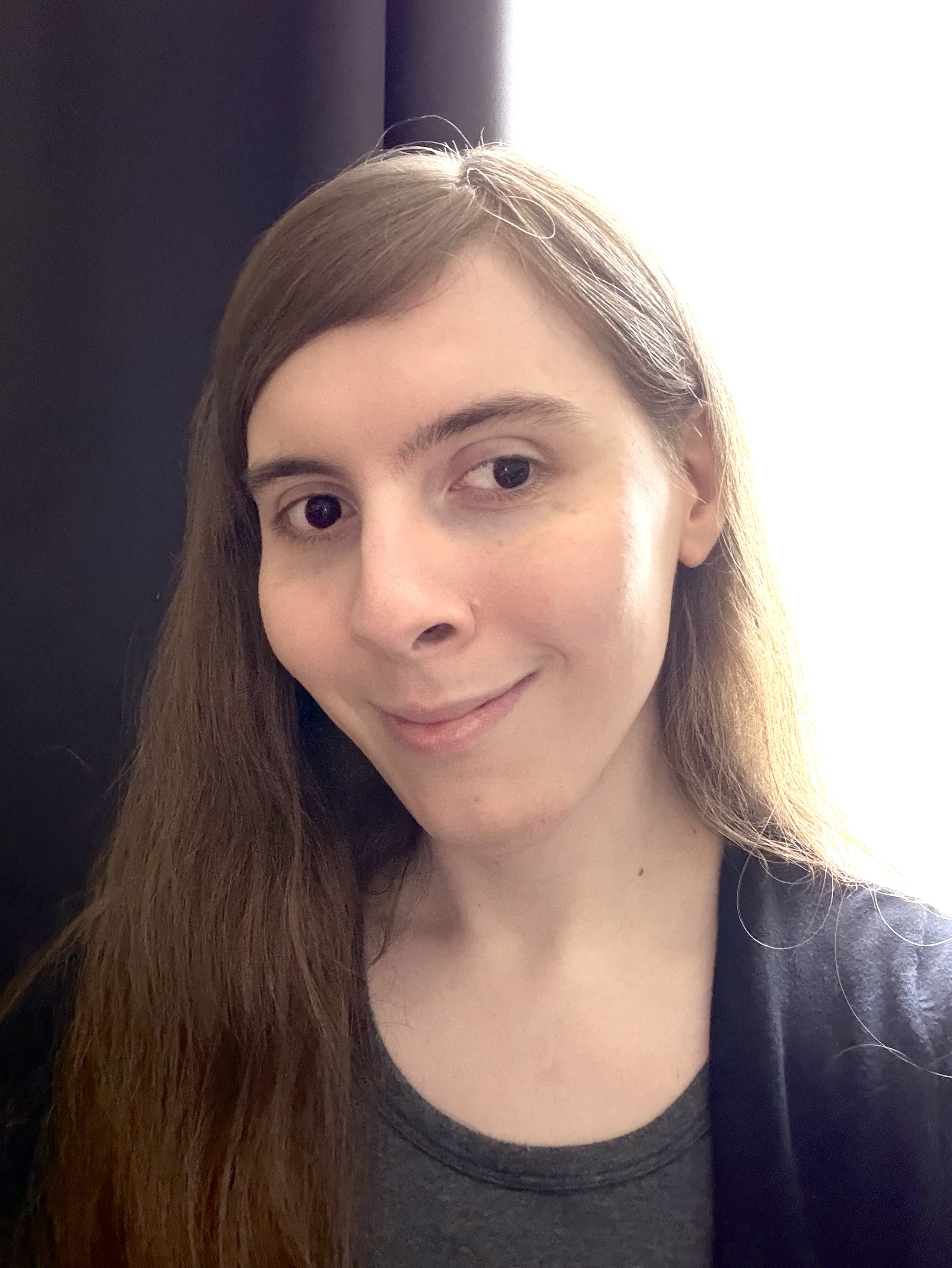 Photo of me! A selfie of a white trans woman with long dark blonde hair and brown eyes wearing a dark scoop-neck tee and a black cardigan smiling and standing in front of a window with dark curtains.