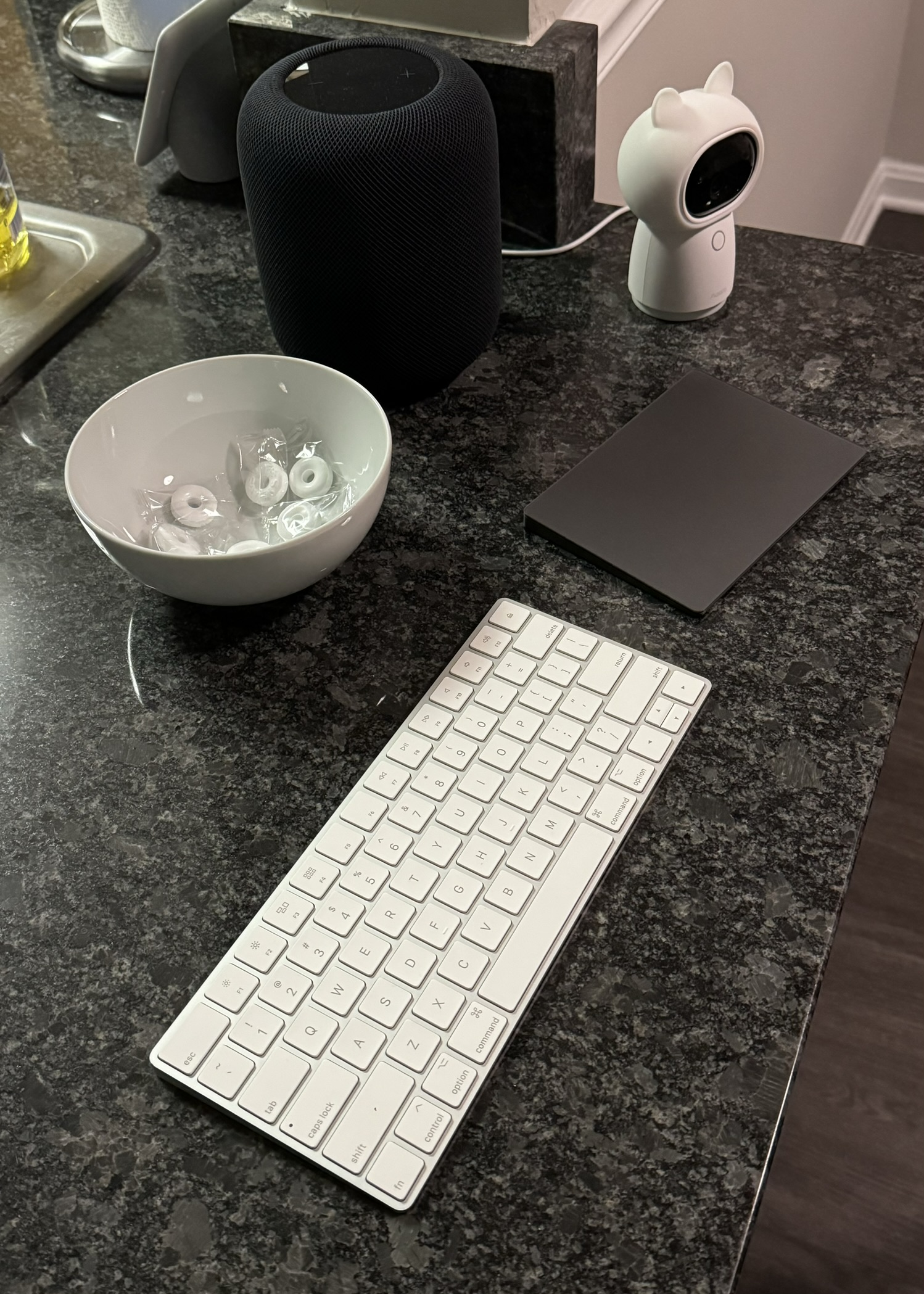 Photo of a white Magic Keyboard and space gray Magic Trackpad sit on a kitchen counter in front of a bowl of Life Savers and a HomePod and next to an Aqara G3 camera.